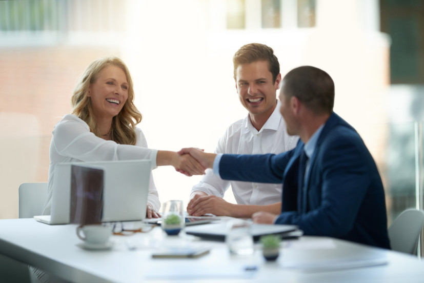 Une commerciale souriante et confiante conclut une vente avec deux clients assis en face d'elle. Ils sont réunis autour d'une table dans un bureau bien éclairé. La commerciale serre la main de l'un des prospects, montrant la finalisation de l’accord.