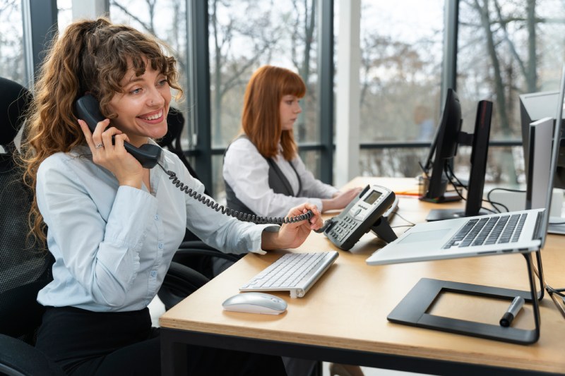 Une commerciale souriante et confiante en train de réaliser une prospection téléphonique dans un bureau moderne et bien équipé. Elle tient un téléphone à la main; un ordinateur devant elle. Son expression radieuse reflète son professionnalisme et son assurance dans ses activités commerciales.