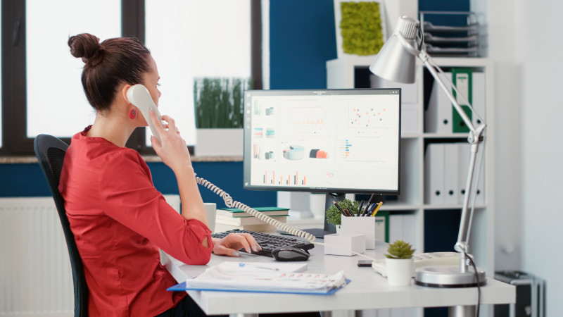 Une commerciale engagée dans une prospection téléphonique, démontrant un haut niveau de concentration. Elle tient un téléphone à la main tout en analysant attentivement les données affichées sur l'écran de l'ordinateur devant elle. Le bureau, baigné de lumière naturelle, est aménagé de manière moderne avec des équipements informatiques sophistiqués.