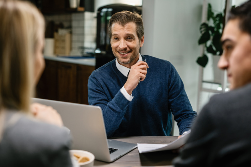 Un vendeur souriant et confiant interagissant avec deux personnes dans un bureau bien éclairé. Son expression faciale démontre assurance et engagement. La scène reflète une communication professionnelle et positive.